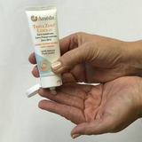 A person's hand holds a small tube of Ameda Triple Zero Lanolin, which is slightly tilted downwards, dispensing a small amount of cream into the palm of the other hand. The tube is primarily white with light blue and orange accents, featuring text that reads "TRIPLE ZERO LANOLIN," along with claims of "Zero Additives, Zero Preservatives, Zero BPA" and "Helps soothe tender, sore nipples." The background is plain and light-colored.