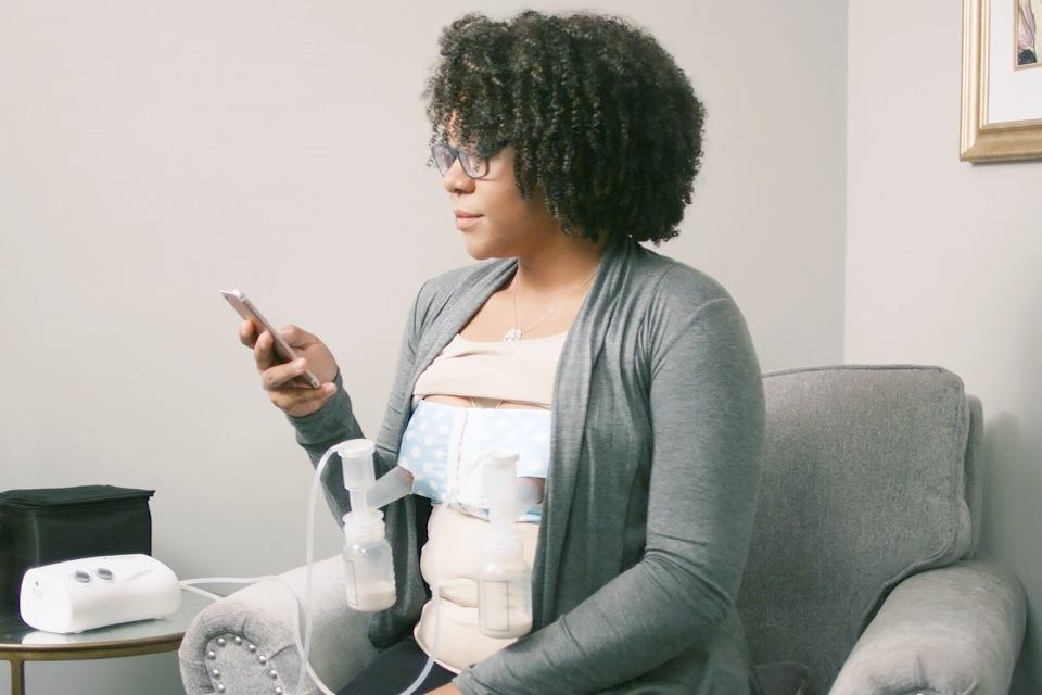 A woman with curly hair sits in a gray chair, wearing a light top and a fitted gray cardigan. She is holding a smartphone in her right hand, looking at the screen. Attached to her chest is a breast pump, connected by tubes to two bottles. A small black bag and a white electronic breast pump are on a side table beside her. The background is a muted color, with a framed piece of art visible on the wall to her right.