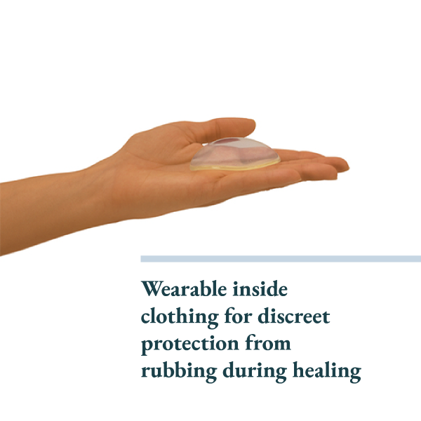 A human hand is shown holding a transparent, dome-shaped item, which appears to be made of soft, flexible material. The item is designed to fit comfortably inside clothing, providing discreet protection against rubbing during the healing process. Beneath the image, there is text that reads, "Wearable inside clothing for discreet protection from rubbing during healing." The background is plain, enhancing the focus on the hand and the item being held.