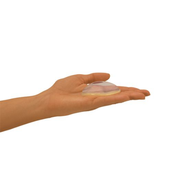 A person’s hand is holding a translucent, dome-shaped object that resembles a silicone gel implant. The hand is facing upwards, with fingers slightly curled around the object while the palm is open. The background is plain and white, emphasizing the hand and the object.