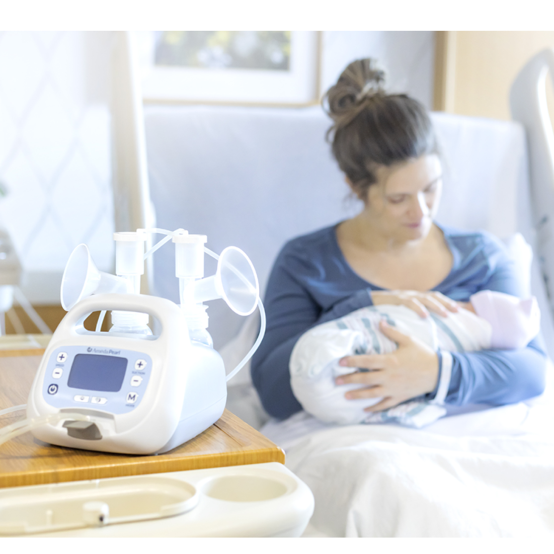 A woman dressed in a long-sleeve blue top sits on a hospital bed, cradling a newborn baby wrapped in a striped cloth. She appears to be gazing down at the baby with a gentle expression. Next to her, on a wooden bedside table, there is a breast pump with two breast shields placed on top, and a digital display with buttons. The background features a light-colored wall with a framed picture and a hospital privacy curtain to the side.