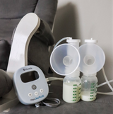 A compact breast pump sits on a gray surface next to two clear plastic bottles, each partially filled with milk and marked with measurement lines. The pump is gray with a digital display and buttons, labeled "Ameda Mya Joy PLUS." There are two attached breast shields resting above the bottles, connected by tubing. In the background, a light gray recliner is slightly visible, adding to a comfortable setting.