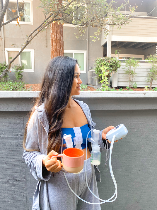 A woman with long, dark hair stands sideways against a gray wall, looking to her left. She wears a gray robe over a blue top and light gray pants. In her left hand, she holds a pink and beige cup, and in her right hand, she holds a small breast pump with tubes attached. The background features a tree and a building with visible windows, hinting at an outdoor setting.